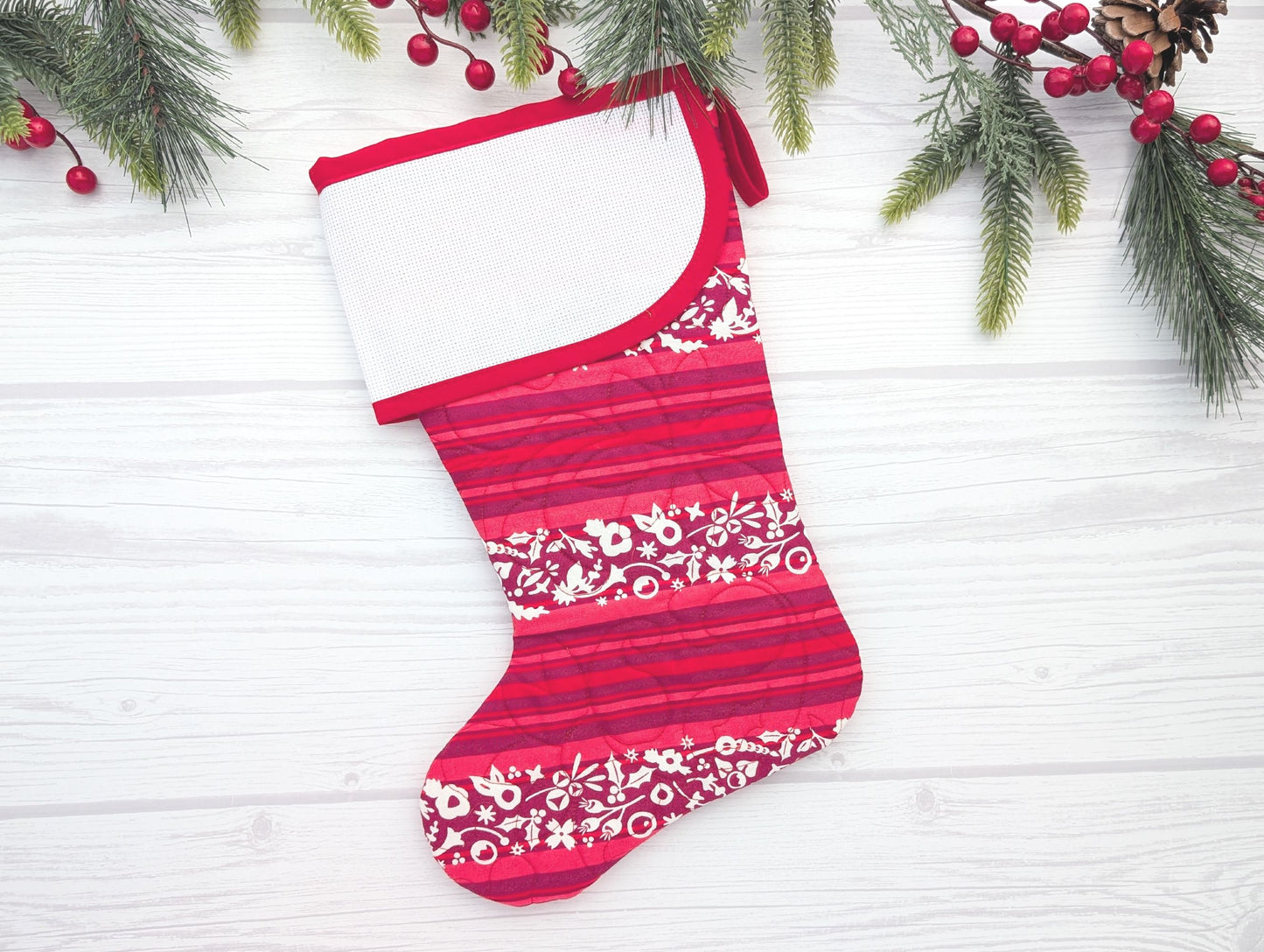 Red and white striped Christmas stocking with floral patterns on a white wooden background with greenery and berries.