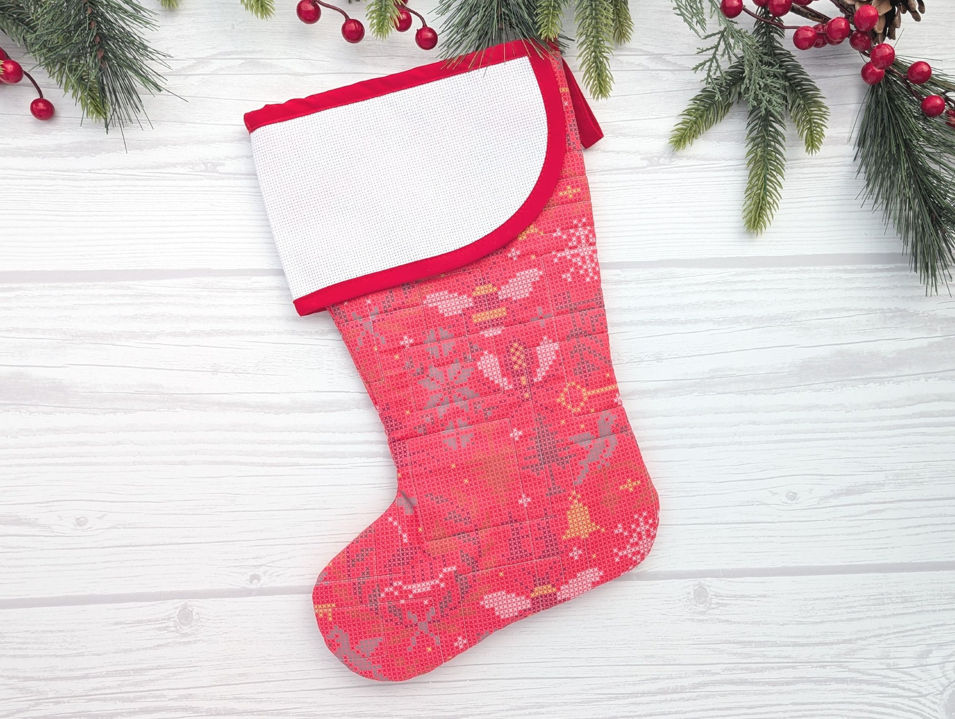 Red Christmas stocking with white trim on a light wooden surface with greenery and berries.
